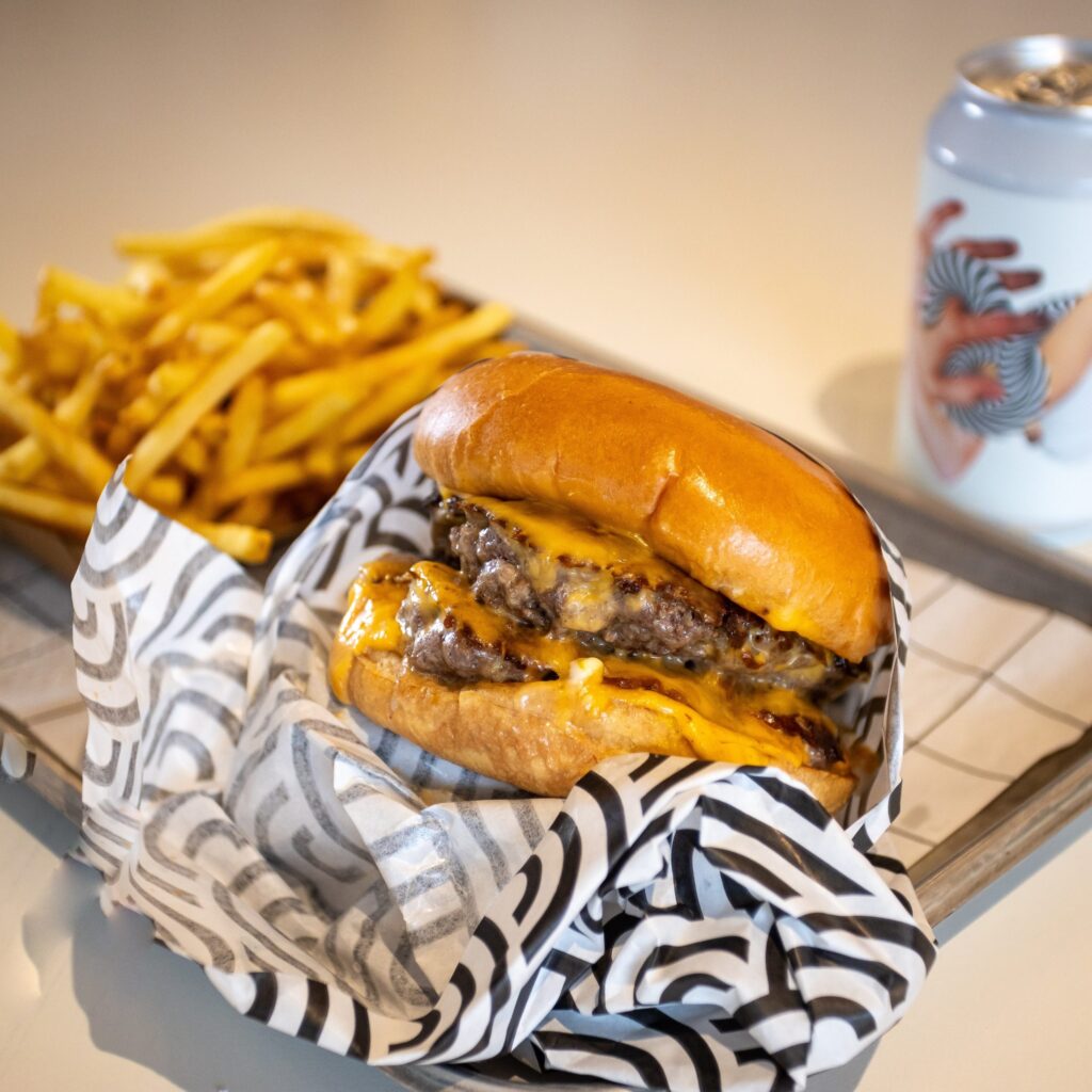 Cheeseburger with chips and a drink served on a tray, wrapped in patterned paper. Indulgent fast food meal.