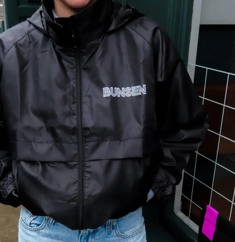 Person wearing a black BUNSEN jacket with hands in pockets, standing against a dark tiled surface.